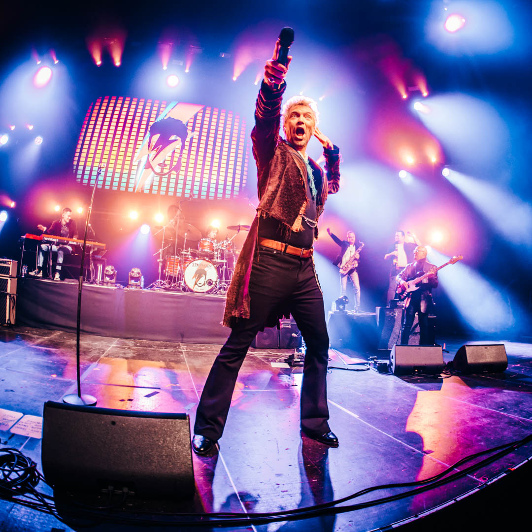 Zanger in glitterkostuum met opgeheven arm op concertpodium in Starman Bowie tribute.