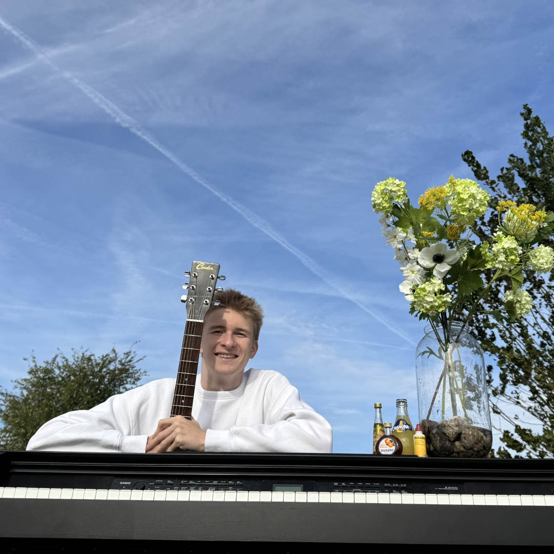Stijn de Groot kijkt over piano met bloemen onder blauwe lucht in cabaretvoorstelling.