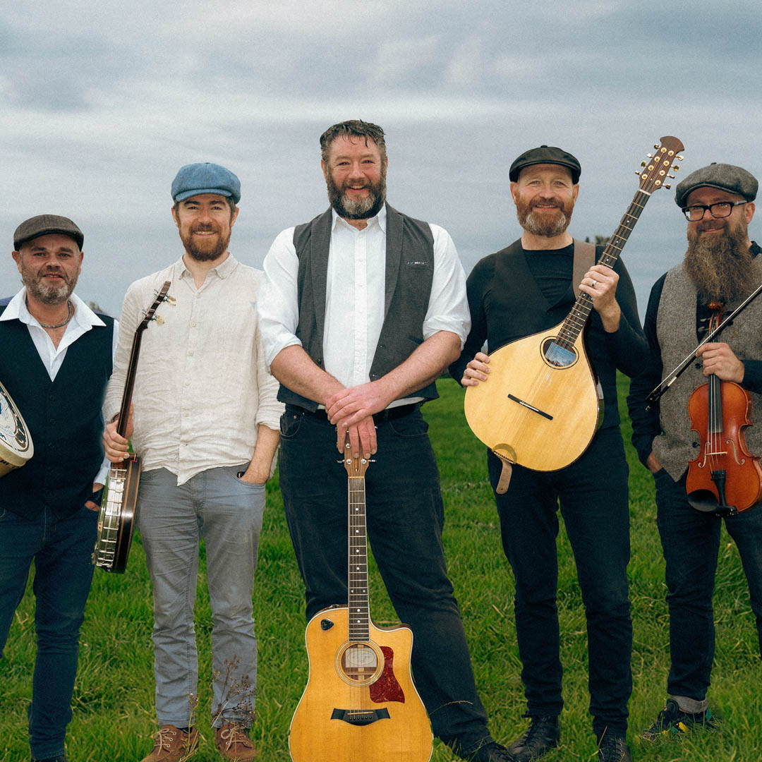 Drie muzikanten van The Dubliners Experience met akoestische instrumenten in een landschap.