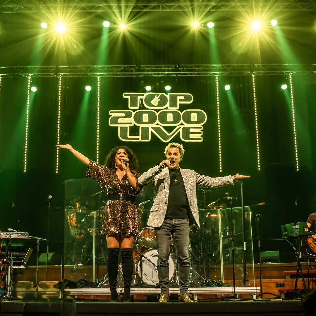 Zangers staan op podium in Top 2000 Live onder groene spotlights met liveband.