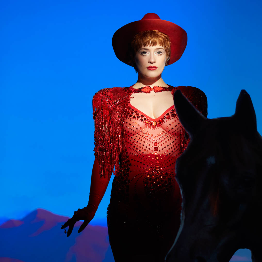 Valentina Tóth in rode outfit met hoed voor blauwe achtergrond in Revue Revanche.
