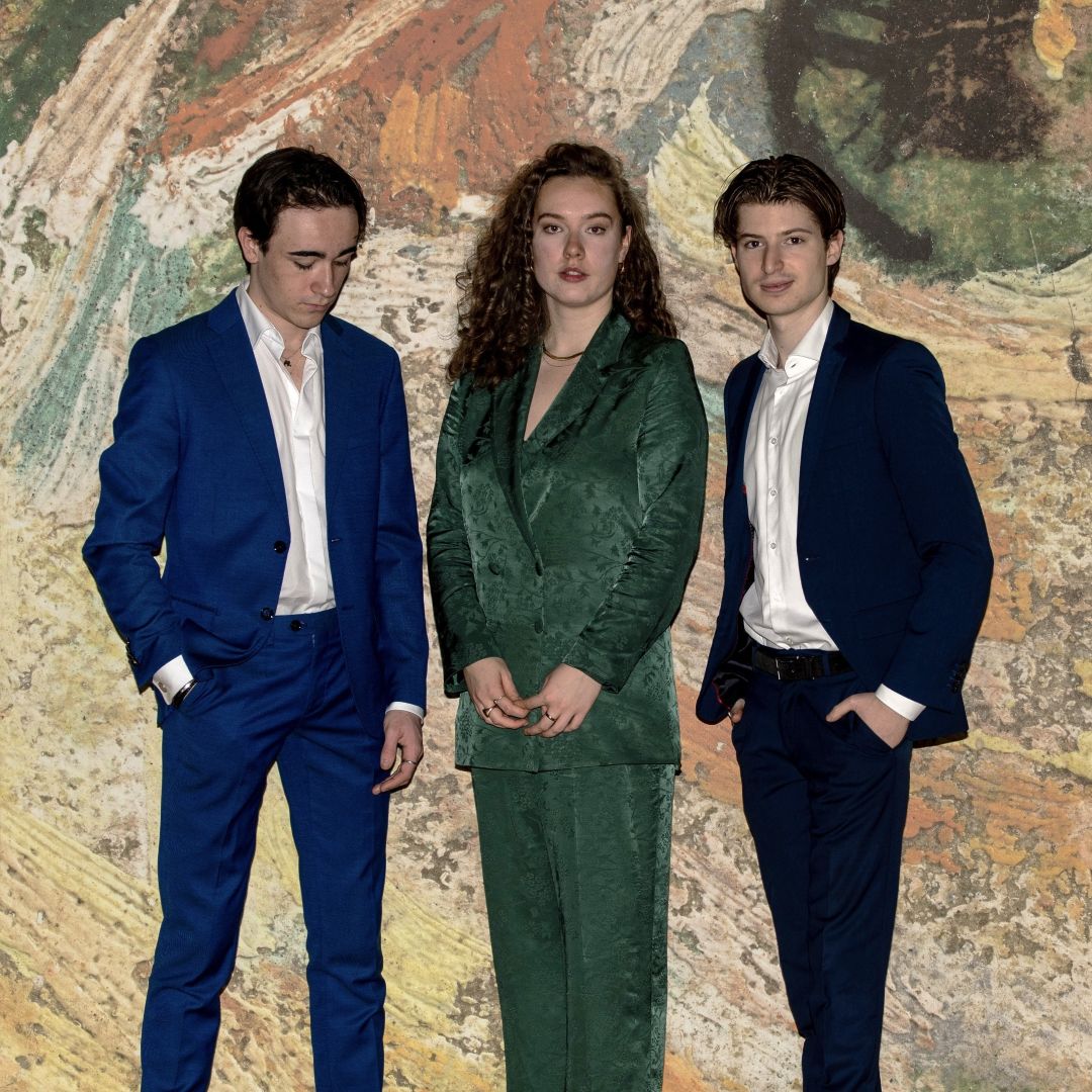 Van Gogh Trio poseert voor geschilderde achtergrond, twee mannen in blauw pak en vrouw in groen pak.