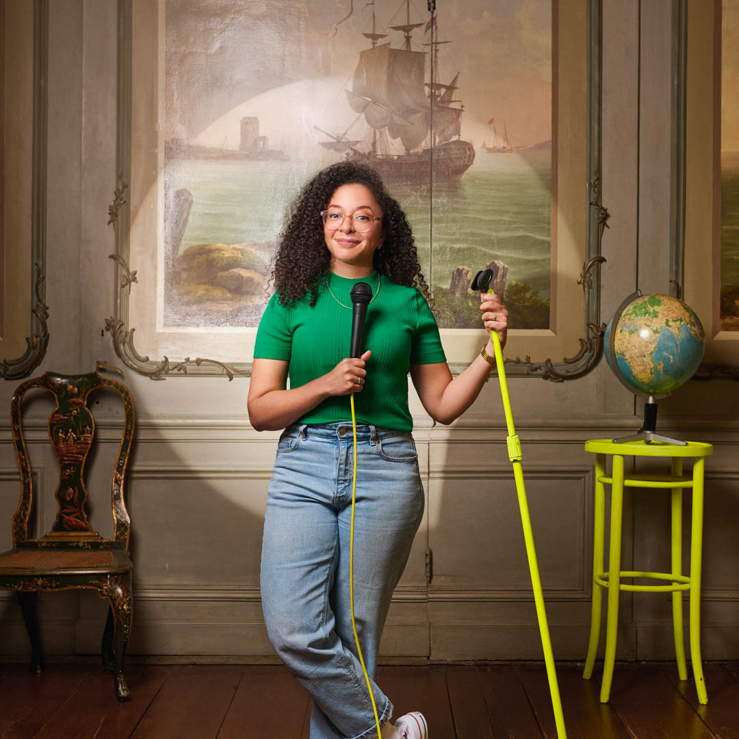 Wina Ricardo staat met microfoon in Wina's Wereld naast felgele kruk op sfeervol decor.