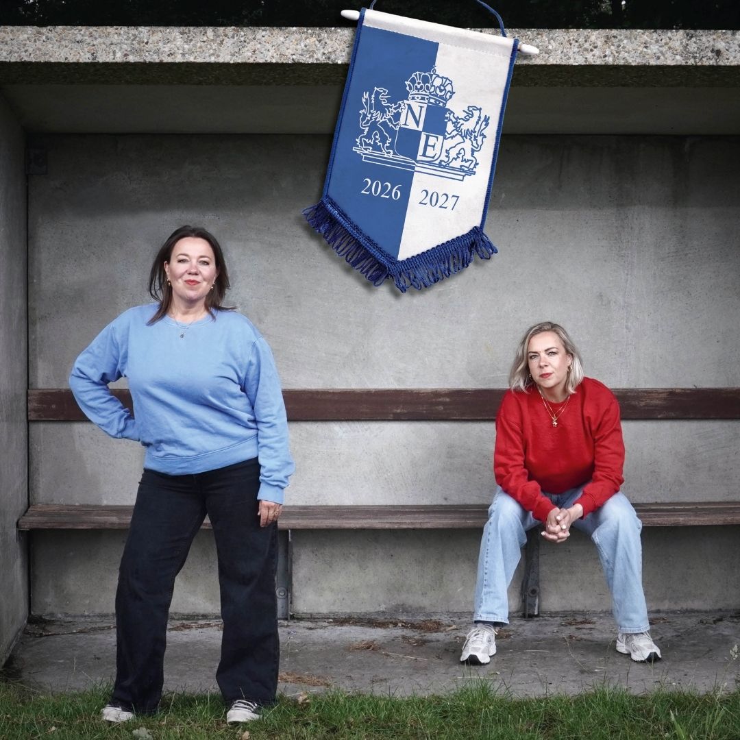 Esther Groenenbergen staat links in een blauwe trui. Zij heeft haar arm in haar zij. Rechts zit Nynke de Jong in haar rode trui op een bankje. In hun midden hangt een vlag.
