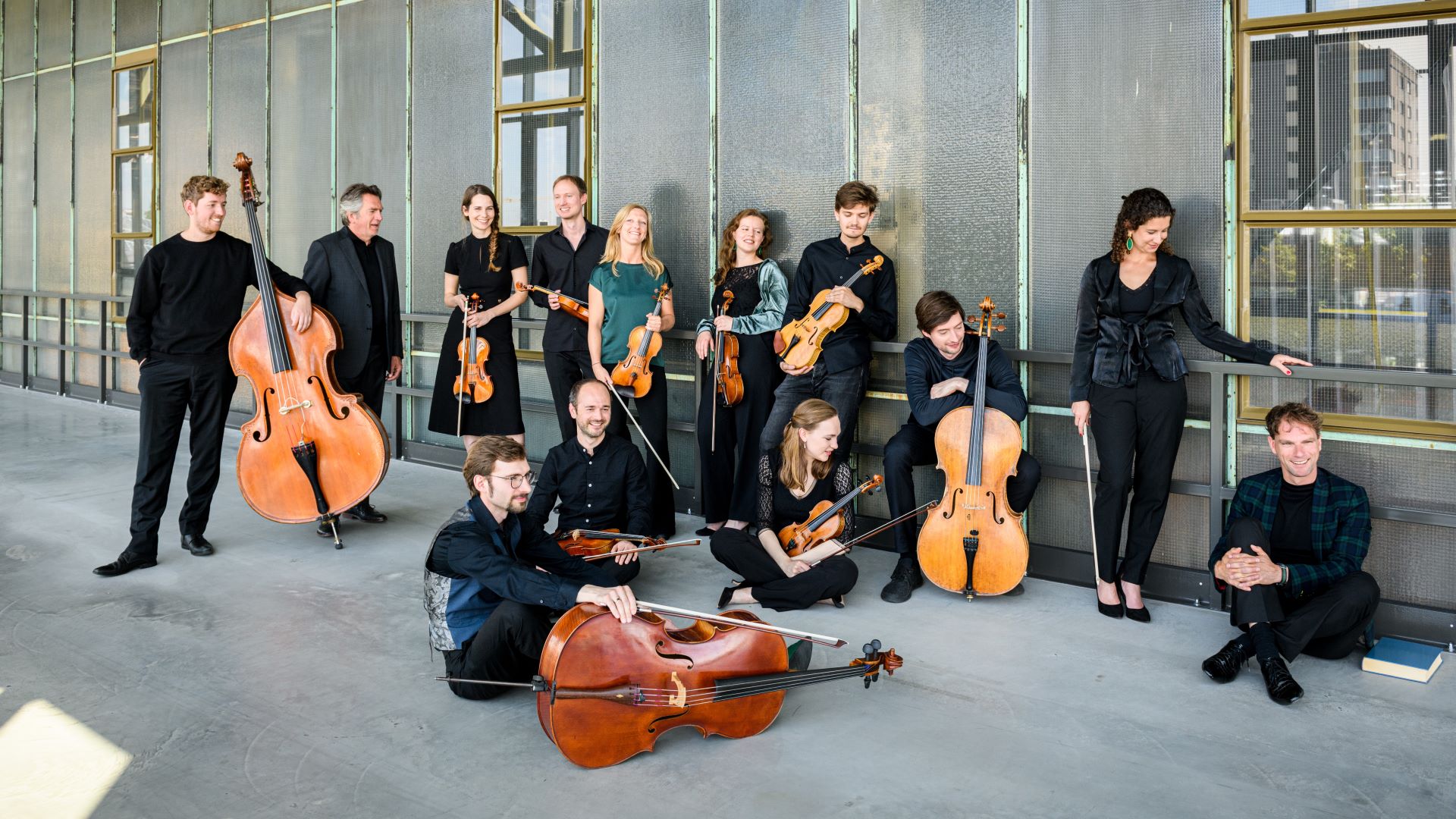 Een groep muzikanten staat en zit langs een hoge glazen wand van een modern gebouw.
