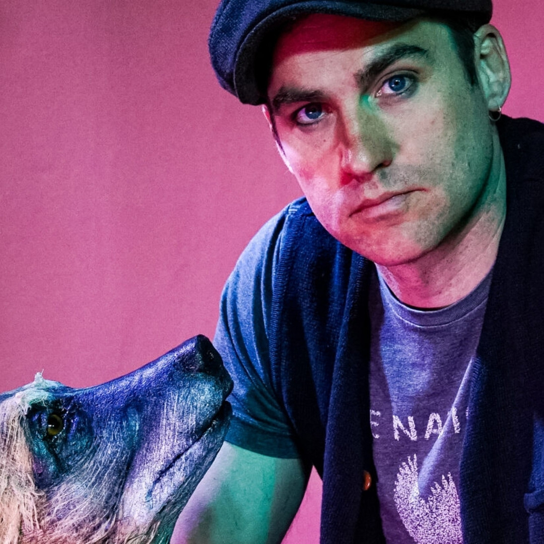 Man met pet kijkt strak in de camera, naast hem een grote hondenpop in blauw-groen theaterlicht.