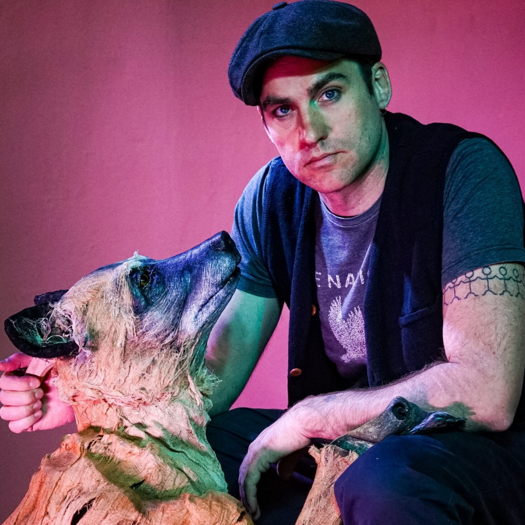 Man met pet kijkt strak in de camera, naast hem een grote hondenpop in blauw-groen theaterlicht.