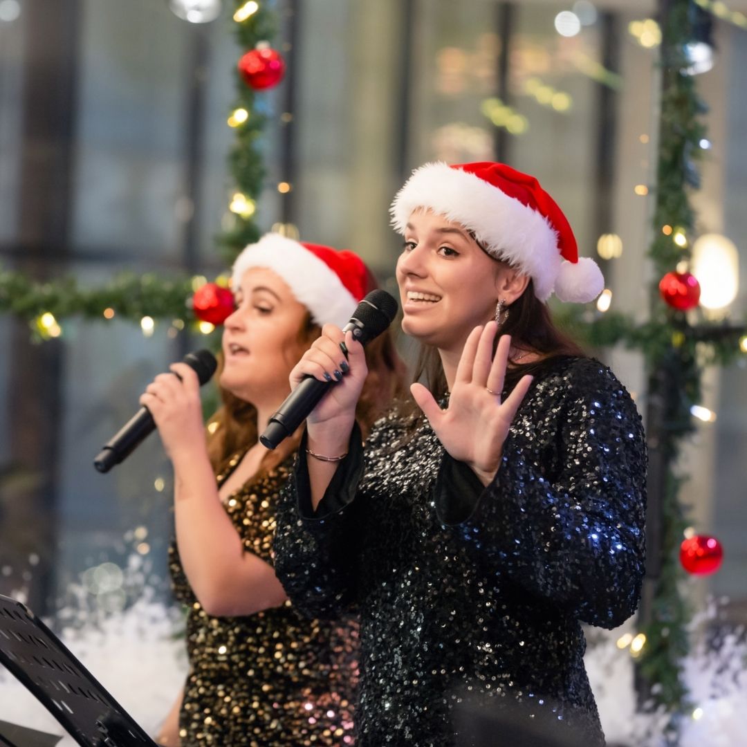 twee vrouwen zingen met een microfoon en hebben een kerstmuts op.