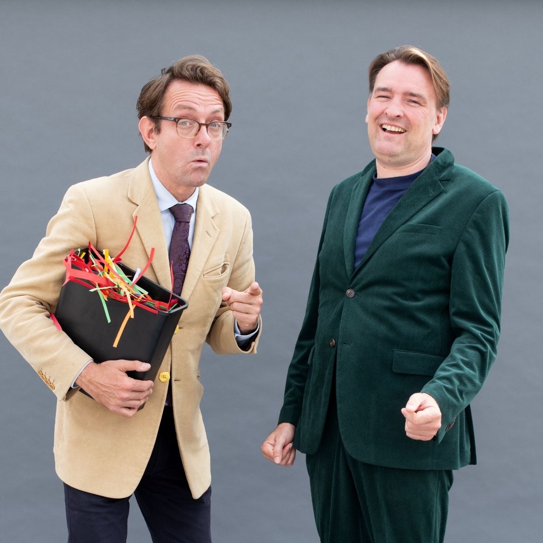 Een man met een bak vol snippers wijst naar de camera terwijl zijn medespeler lacht tijdens Woordgrapf.